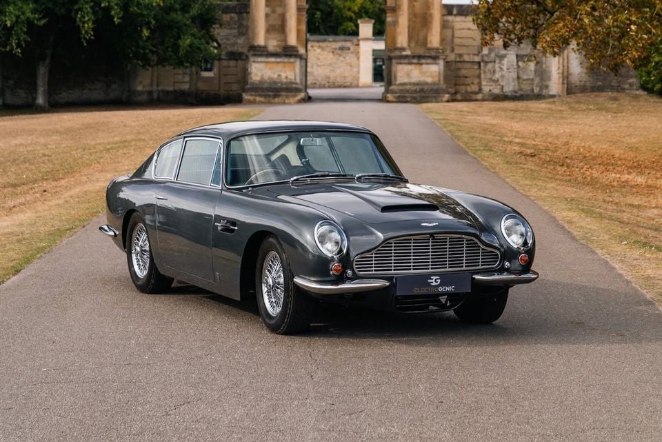 A grey Aston Martin DB6 coupe parked on a driveway in front of a grand stone building, seen from the front-side