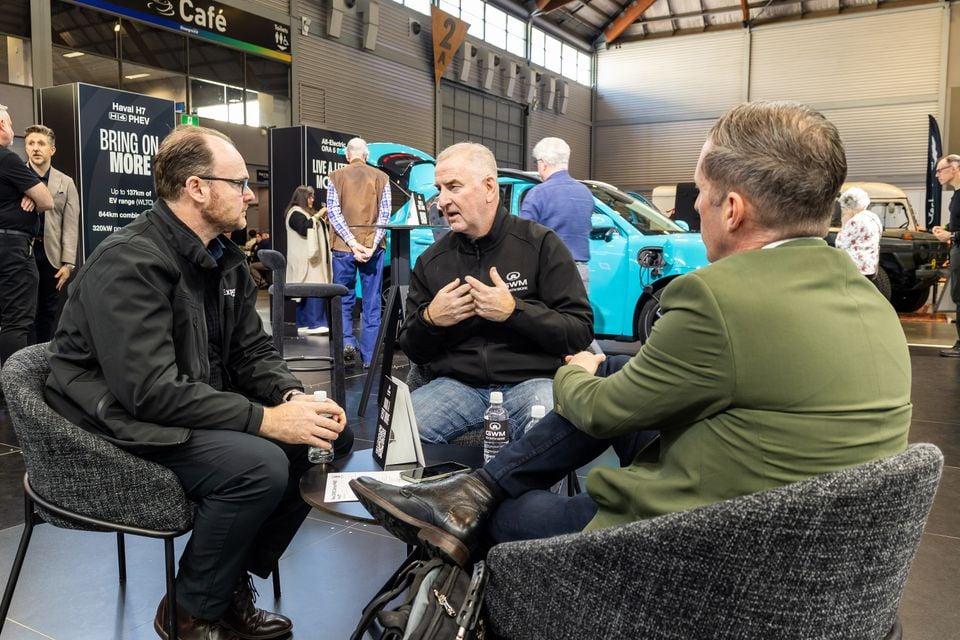 Three men sit talking at a GWM display stand during an electric vehicle event, with a turquoise electric car in the background