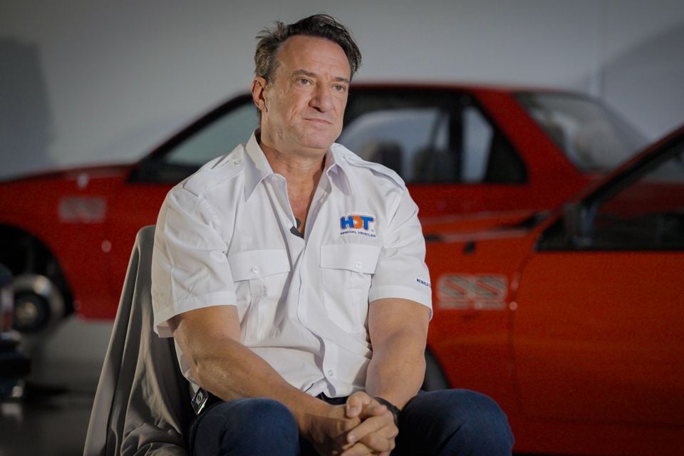 A man in a white HDT Special Vehicles shirt seated for an interview in front of red Holden Commodore SS cars