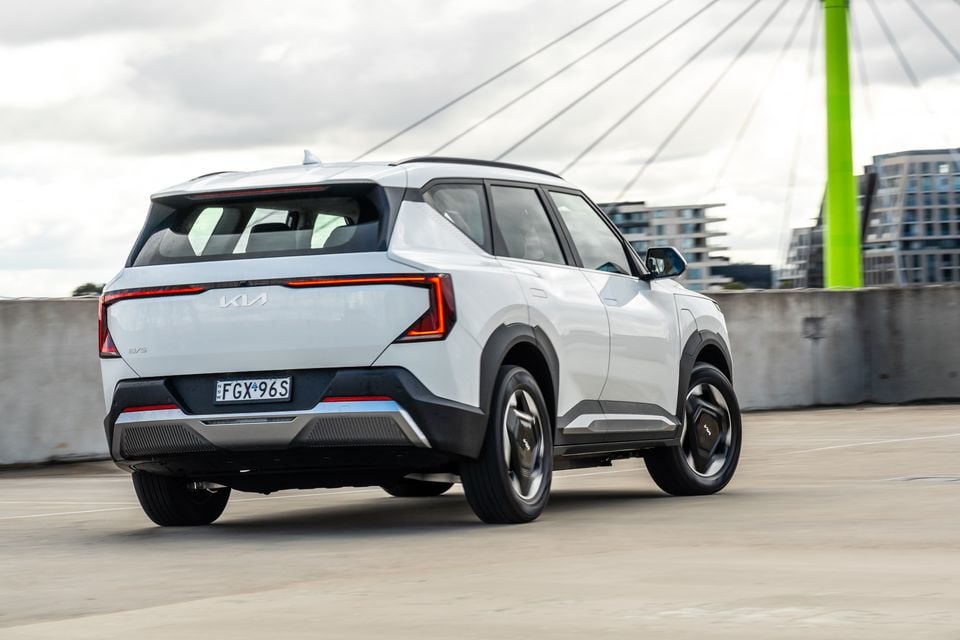 A white Kia EV5 SUV seen from the rear three-quarter angle on a rooftop car park with a city backdrop