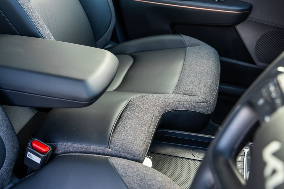 Interior view of a Kia EV5 showing the front centre console area with dark fabric and leather seats, armrest, and seatbelt buckle