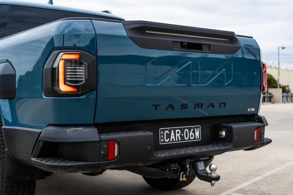 Rear of a teal Kia Tasman X-Pro ute showing the tailgate with embossed Kia logo and Tasman lettering, LED tail lights, and a tow bar fitted