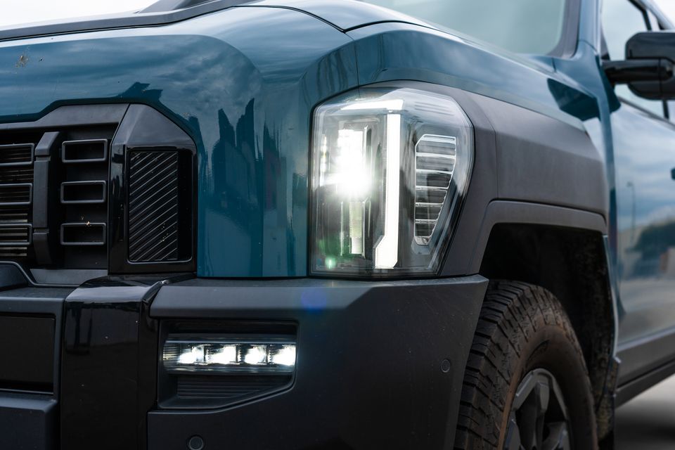 Close-up of the LED headlight and front corner of a teal Kia Tasman X-Pro ute, with fog lights illuminated and off-road tyres visible