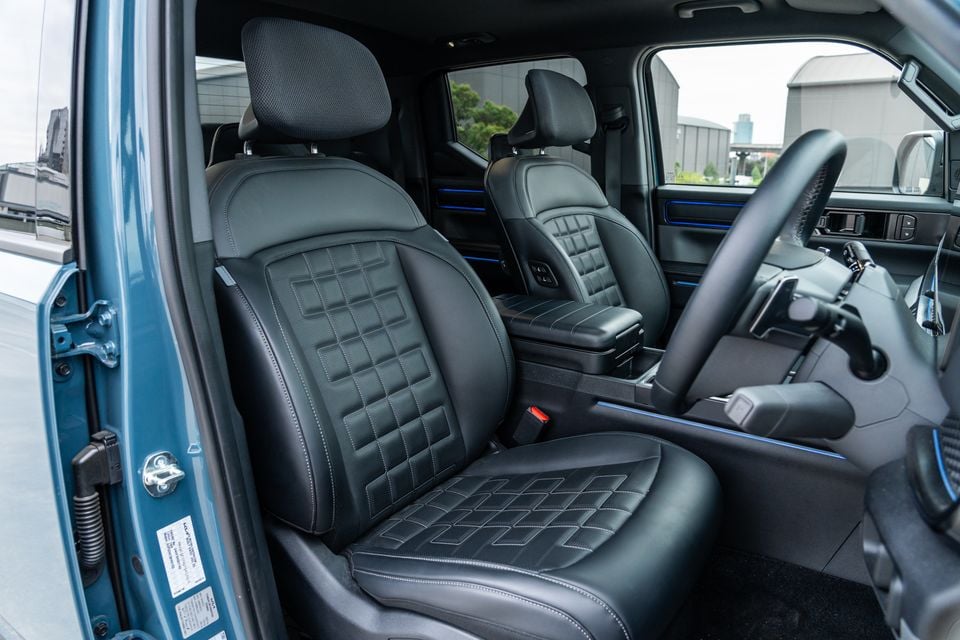 Black leather-trimmed front seats with quilted stitching inside a Kia Tasman X-Pro, viewed through the open driver's door with blue ambient lighting accents visible