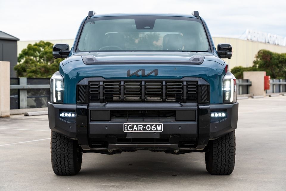 Front view of a teal Kia Tasman X-Pro ute parked in a car park, showing its bold grille, LED headlights and bull bar bumper
