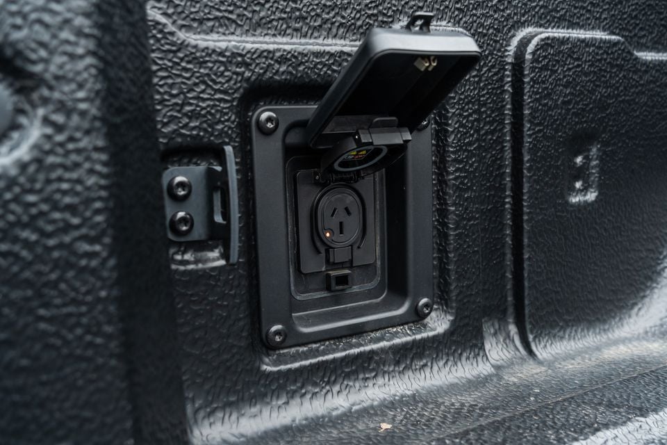 An Australian-style three-pin power outlet built into the textured cargo area of a Kia Tasman X-Pro ute, with the protective cover open