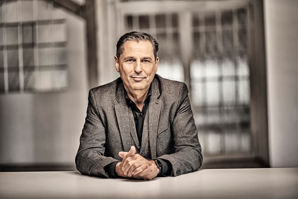 A man in a grey blazer sits at a table with his hands clasped, smiling at the camera in an office setting