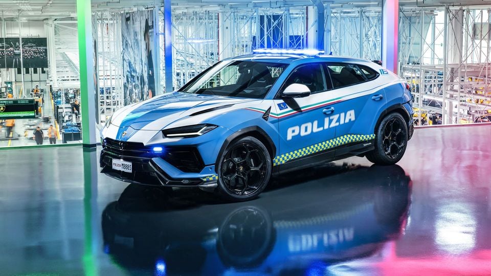 A Lamborghini Urus SUV in Italian Polizia livery, finished in blue and white with a yellow checkered stripe, photographed from the front-side inside what appears to be the Lamborghini factory.