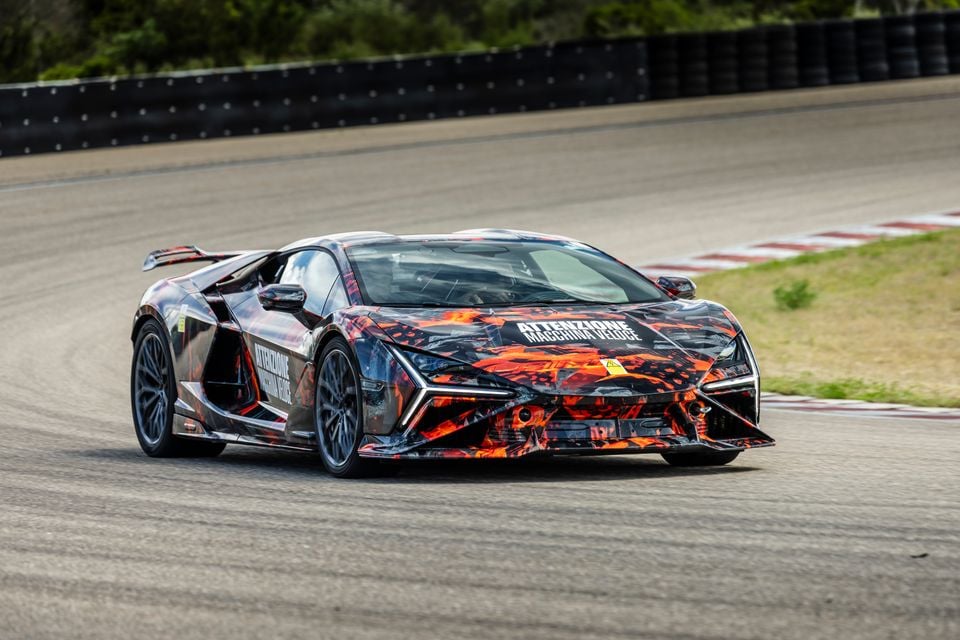 A Lamborghini Revuelto supercar wearing a striking orange and black camouflage wrap, seen from the front-side angle on a race track