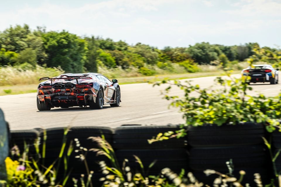 A camouflaged Lamborghini Revuelto SV prototype seen from the rear while driving on a test track, with a second supercar visible ahead in the distance