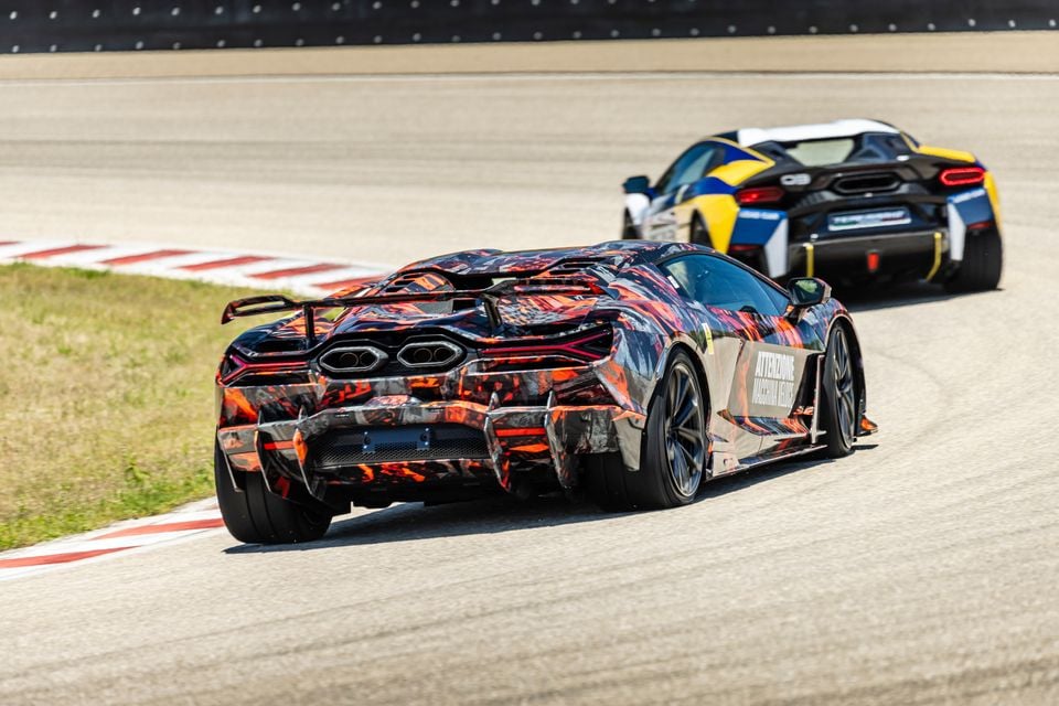 A Lamborghini Revuelto SV prototype in camouflage wrap drives on a race track, seen from the rear, with a second supercar following behind