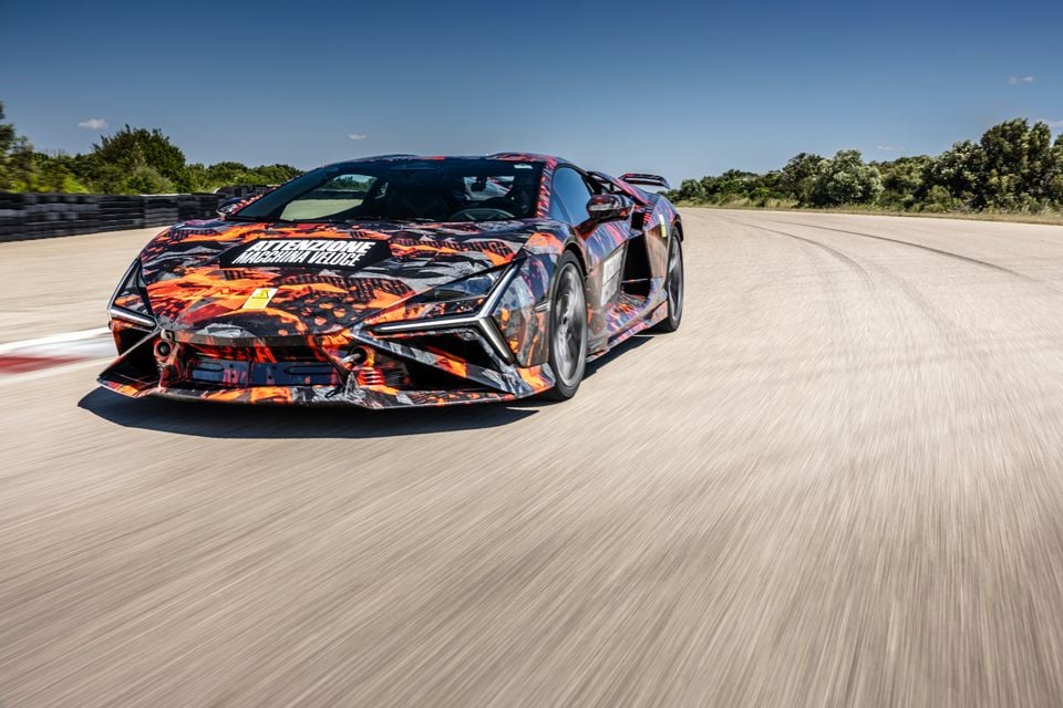 A Lamborghini Revuelto SV prototype in orange and black camouflage wrap driving at speed on a test track, seen from the front side