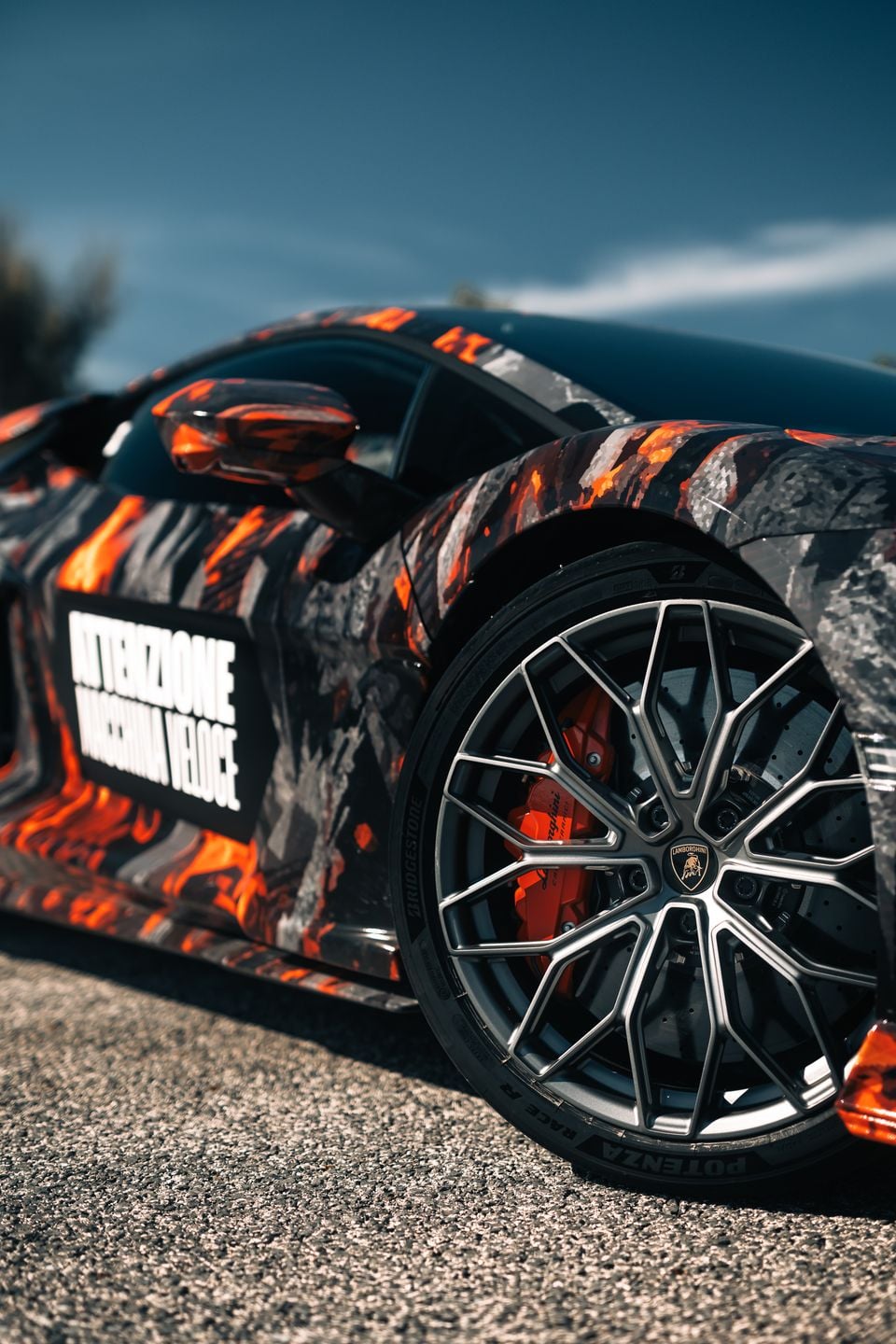 Close-up of a Lamborghini Revuelto supercar with a dramatic lava-themed wrap, showing the rear wheel with orange brake calipers and a Lamborghini badge on the alloy wheel