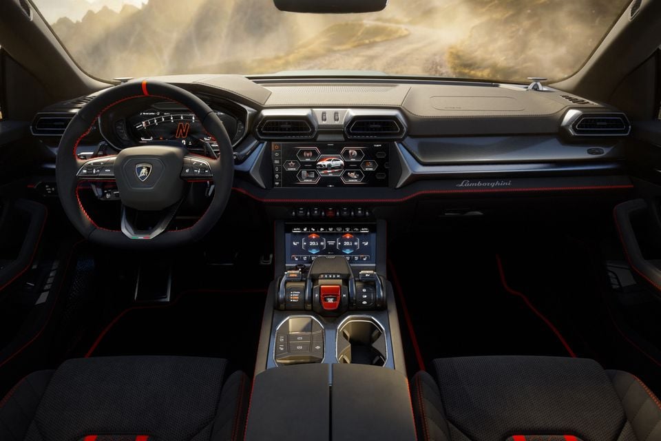 Lamborghini Urus interior showing the dashboard, dual touchscreens, carbon fibre trim, sport steering wheel with red stitching, and centre console with drive mode selector