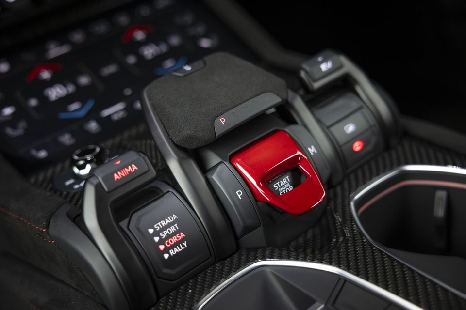Close-up of the Lamborghini Urus centre console showing the ANIMA drive mode selector with Strada, Sport, Corsa and Rally modes, a red Start Engine Stop button, and gear selector controls on a carbon fibre surround