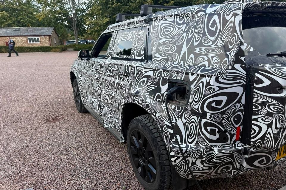 A camouflaged Land Rover Defender prototype showing the side and rear quarter, with a visible charging port, parked on a gravel surface during testing.