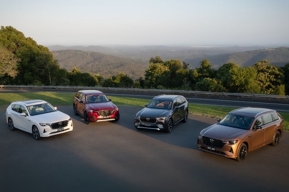 Four Mazda SUVs — including the CX-60, CX-70, CX-80, and CX-90 — parked together on a hilltop road with a scenic Australian landscape in the background.