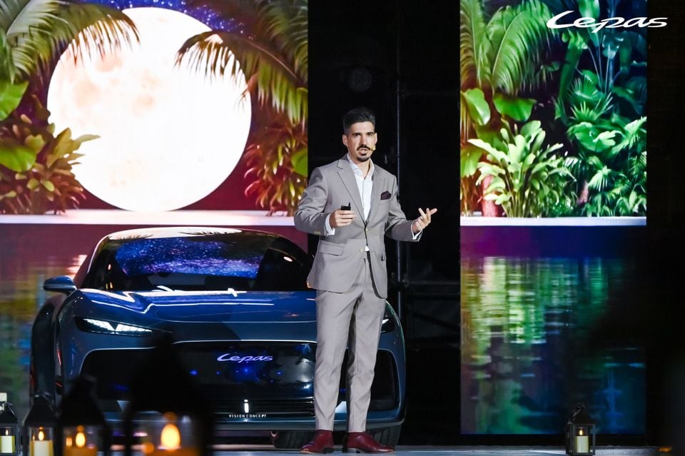 A presenter in a grey suit speaks on stage in front of a dark blue Lepas Vision Concept car at a launch event