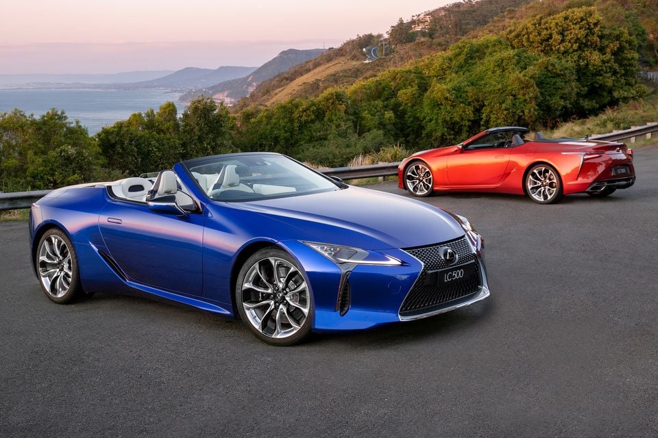 Two Lexus LC 500 convertibles parked on a coastal road, one in blue seen from the front three-quarter angle and one in red seen from the rear, with hills and ocean in the background.