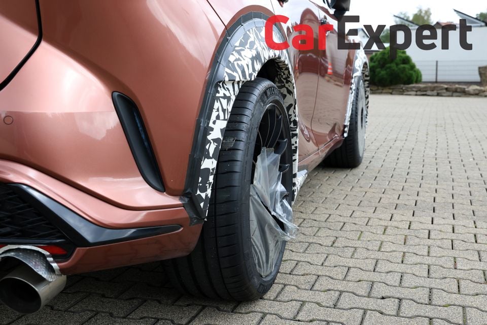Close-up of the rear wheel arch and side body of a camouflaged red Lexus RX Morizo RR prototype, showing wrapped wheel arch and Michelin tyre on a paved surface
