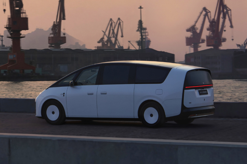 A white Li Auto MEGA electric minivan seen from the side, driving along a waterfront road with industrial cranes in the background at dusk.