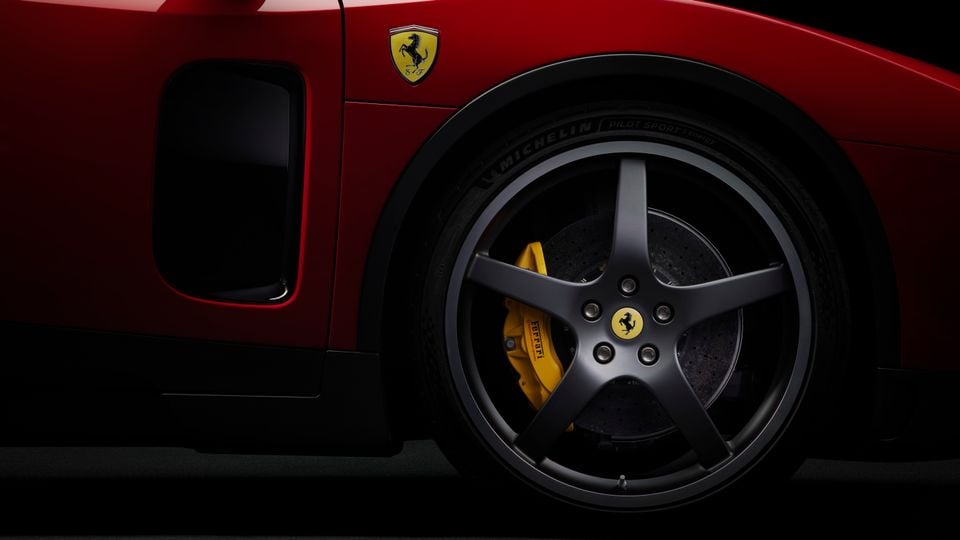 Close-up of a red Ferrari's rear quarter panel showing a five-spoke dark alloy wheel with Michelin Pilot Sport tyre, yellow Ferrari brake caliper, and the Prancing Horse badge on the bodywork