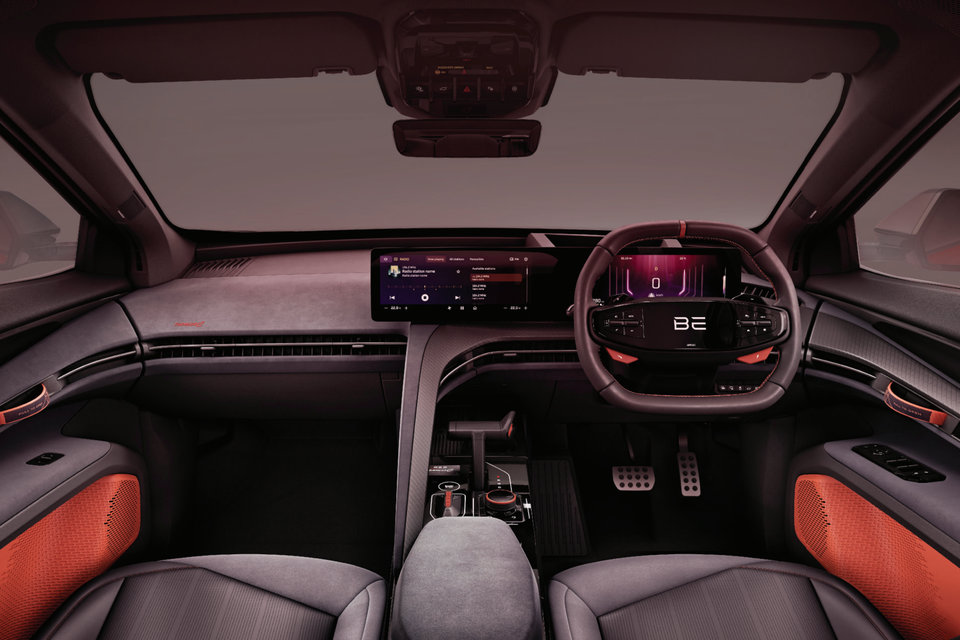 Interior of the Mahindra BE 6 showing the dashboard, wide central infotainment touchscreen, digital instrument cluster, flat-bottom steering wheel, and orange accent door panels with right-hand drive layout