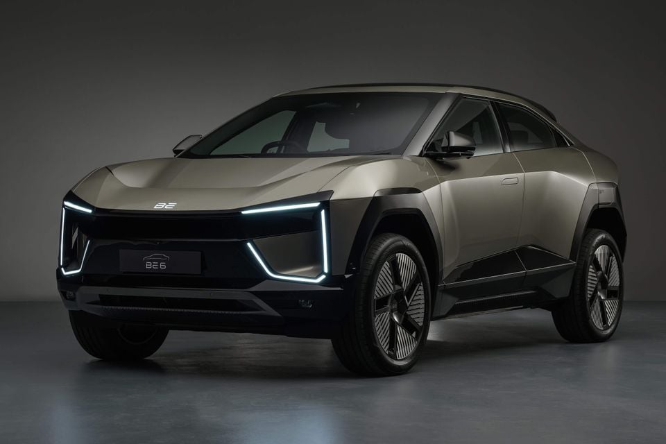 A Mahindra BE 6 electric SUV coupe in gold/bronze, seen from the front-side angle in a studio setting