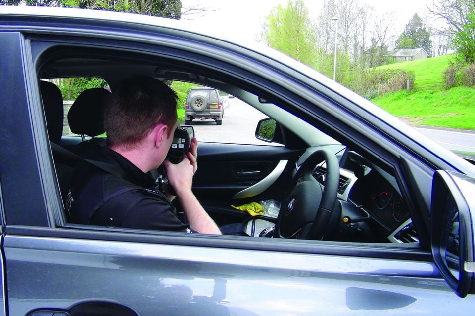 A police officer inside a BMW sedan aims a speed radar gun at traffic on a rural road