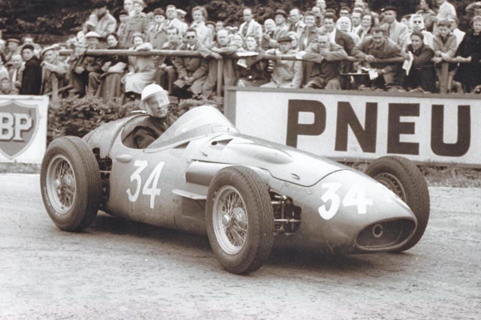 A Maserati 250F racing car number 34 cornering at Spa in a black-and-white photo, with spectators lining the barrier behind