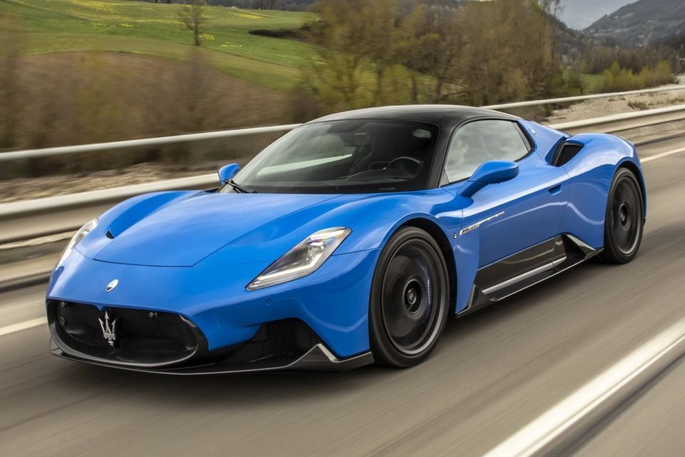 A blue Maserati MC20 supercar with a black roof driving along a country road, seen from the front left