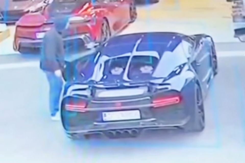 A black Bugatti Chiron seen from above and behind in a car park, with a hooded person at the driver's door, captured on security camera