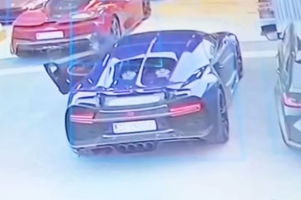 A dark Bugatti Chiron seen from above and behind with its driver's door open, parked beside a red sports car in grainy security camera footage