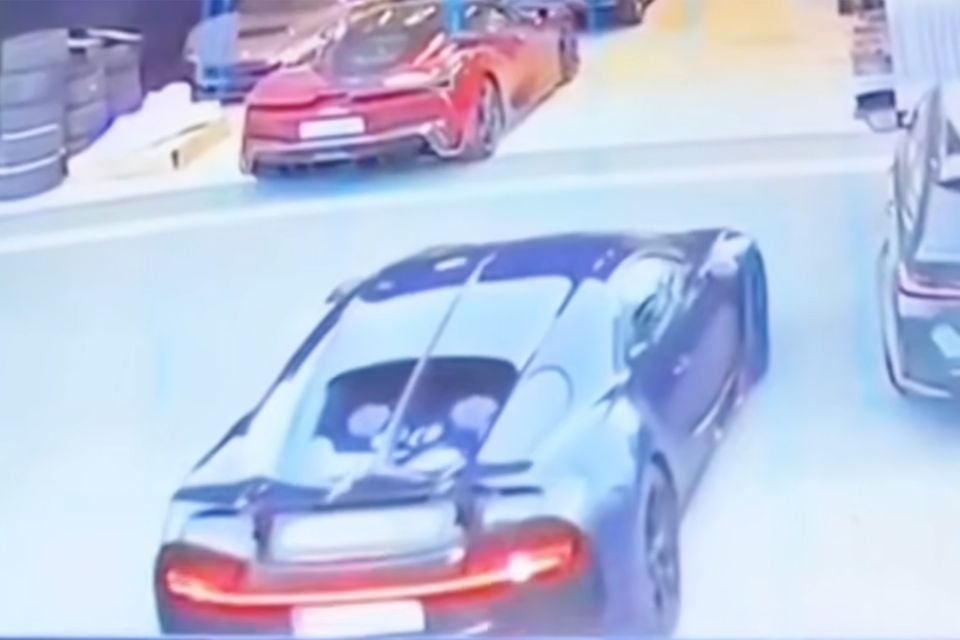 A dark blue Bugatti Chiron seen from above and behind in CCTV-style footage, driving past a red sports car in a garage area