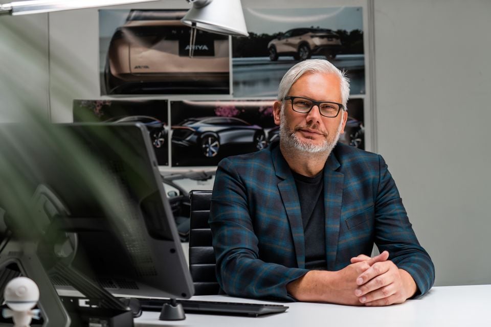 A man with grey hair and glasses sits at a desk in a design studio, with Nissan Ariya design images displayed on the wall behind him.