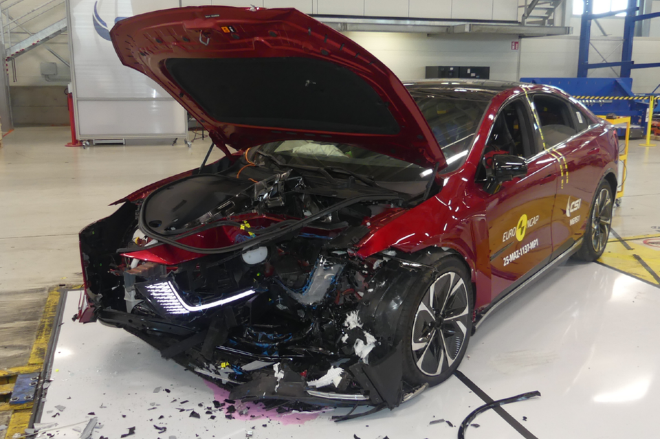A red Mazda 6e sedan after a Euro NCAP frontal crash test, showing severe front-end damage with the bonnet raised and debris on the floor