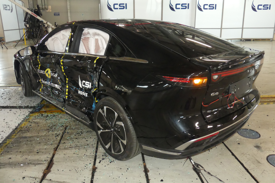 A black Mazda 6e sedan after a side-impact Euro NCAP crash test, showing significant door damage and deployed airbags, photographed from the rear three-quarter angle inside a CSI testing facility.