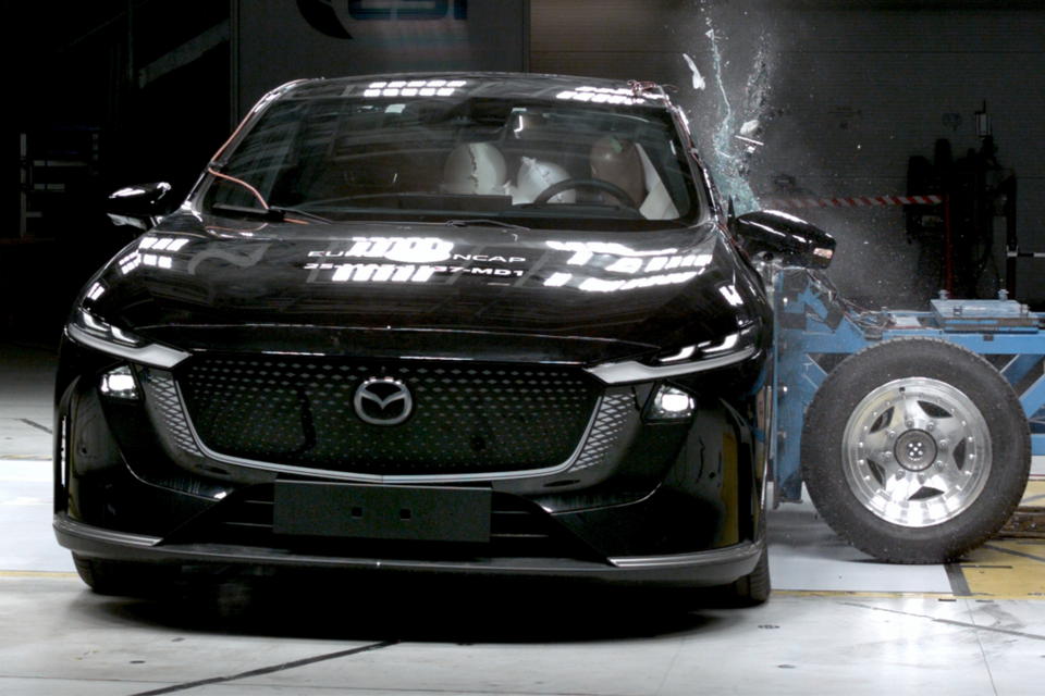 A black Mazda 6e undergoing a side pole crash test with Euro NCAP, seen from the front as debris flies from the passenger side window.