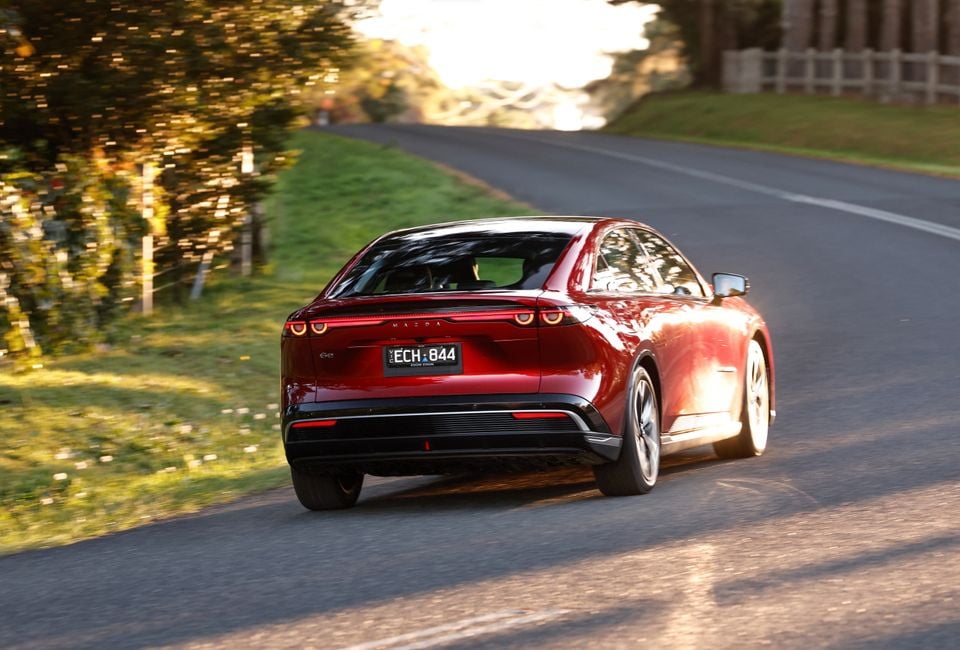 A red Mazda 6e sedan driving on a country road, seen from the rear quarter