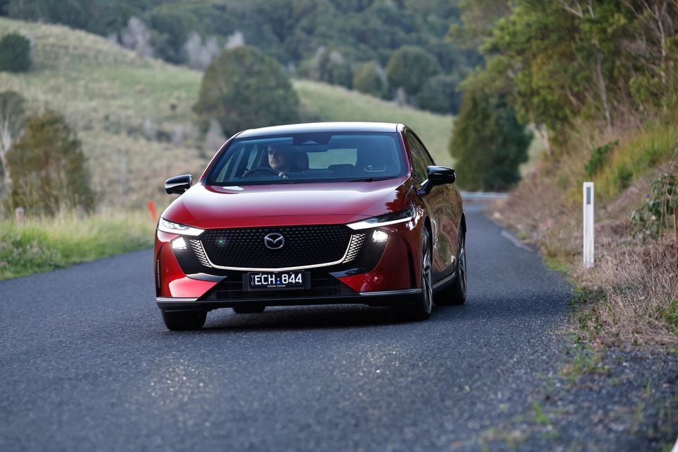 A red Mazda 6e sedan being driven on a rural road, seen head-on.