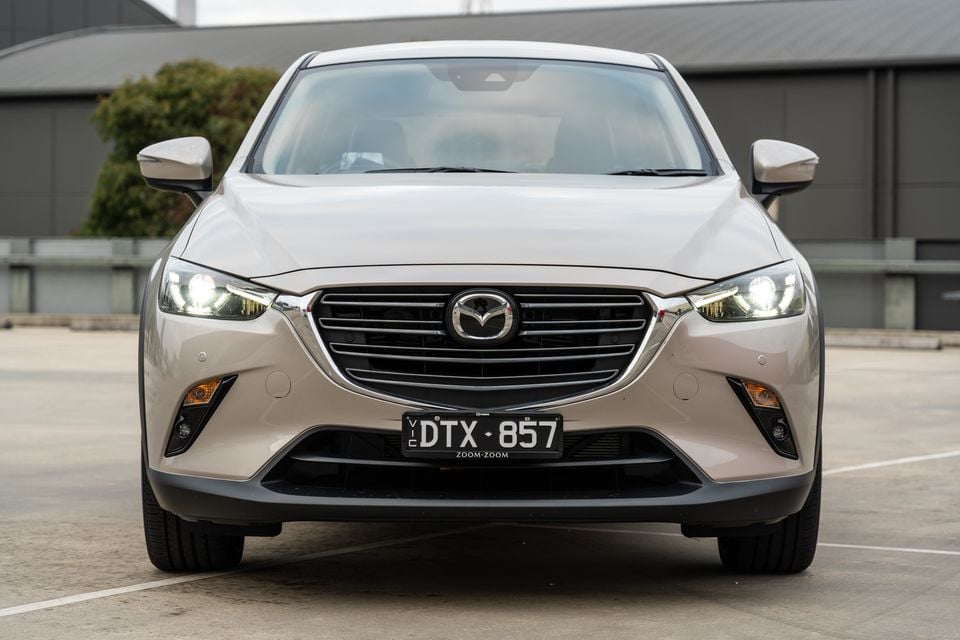 Front view of a beige Mazda CX-3 small SUV parked in a car park with headlights on
