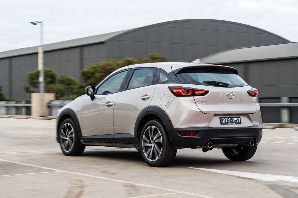 A silver Mazda CX-3 SUV seen from the rear three-quarter angle in a car park, with a large industrial building in the background