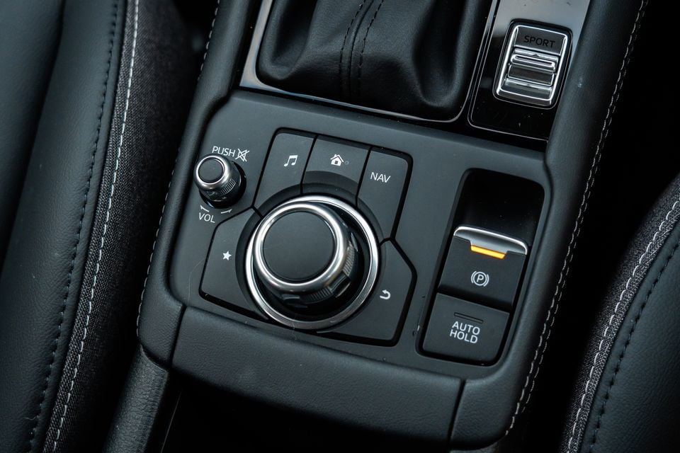 Close-up of the centre console controls in a Mazda CX-3, showing the rotary infotainment dial, volume knob, NAV, Sport mode, electric parking brake, and Auto Hold buttons