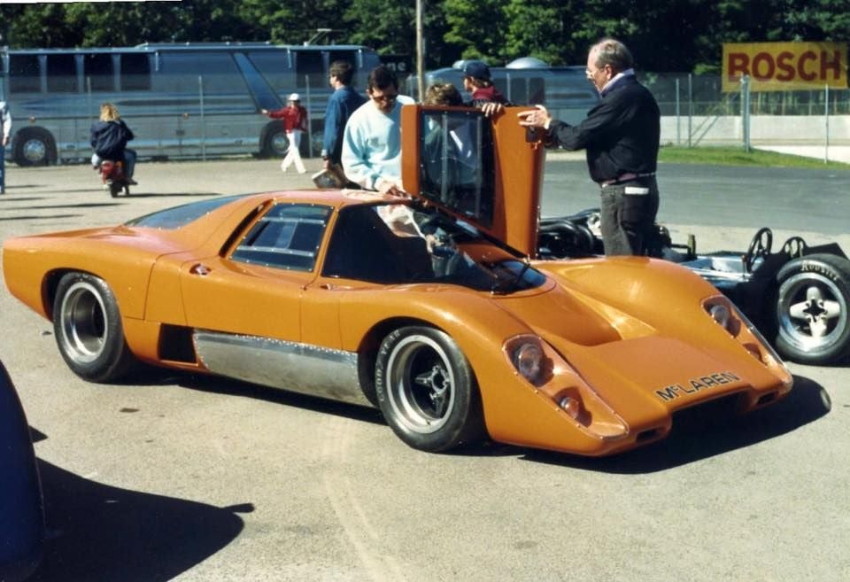 An orange McLaren M6GT sports car with its engine cover open, parked in a paddock area at a racing circuit, with people gathered around it