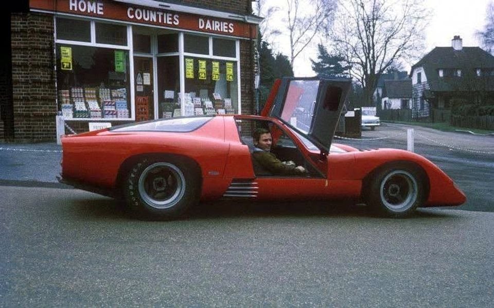 A red McLaren M6GT sports car with its gull-wing door open, seen from the side on a street in front of a Home Counties Dairies shop, with a person seated inside