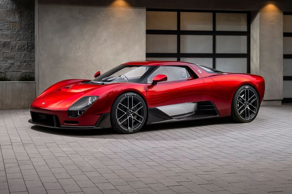 A red McLaren MCL6GT supercar seen from the front-side angle, parked on a brick paved driveway in front of a modern building