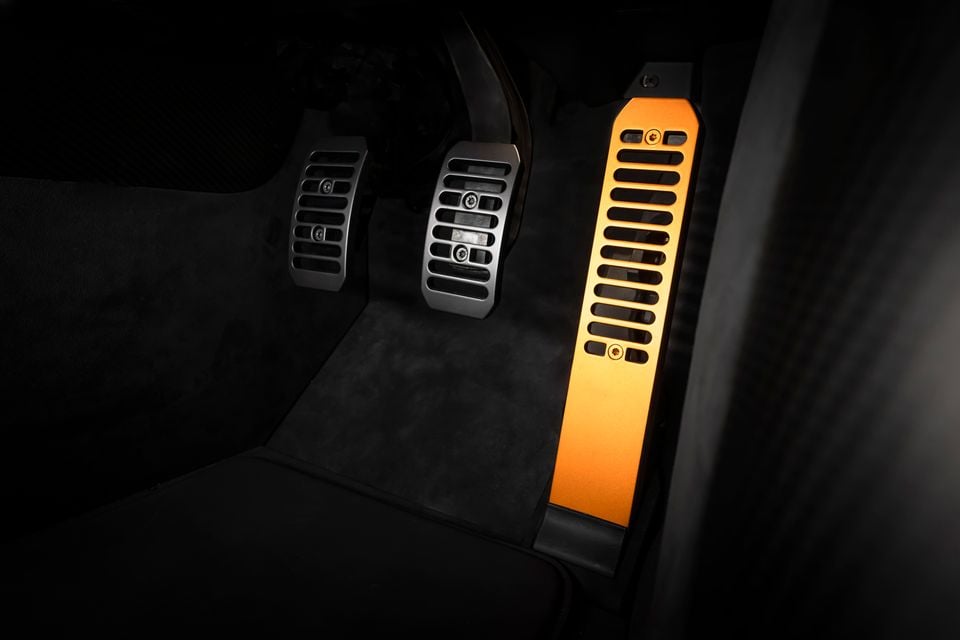 Aluminium sport pedals inside a McLaren, with two silver pedals and a striking orange accelerator pedal on dark carpet