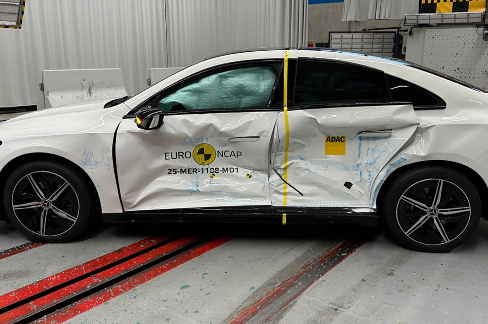 A white Mercedes-Benz CLA seen from the side after a Euro NCAP side-impact crash test, showing significant door deformation and deployed curtain airbags inside a testing facility.