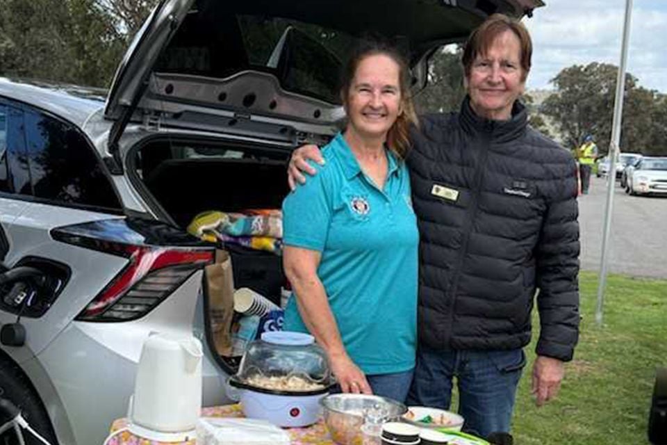 Two people smiling beside a silver MG4 with its tailgate open, using vehicle-to-load power to run a popcorn maker at the Rob Roy Hillclimb