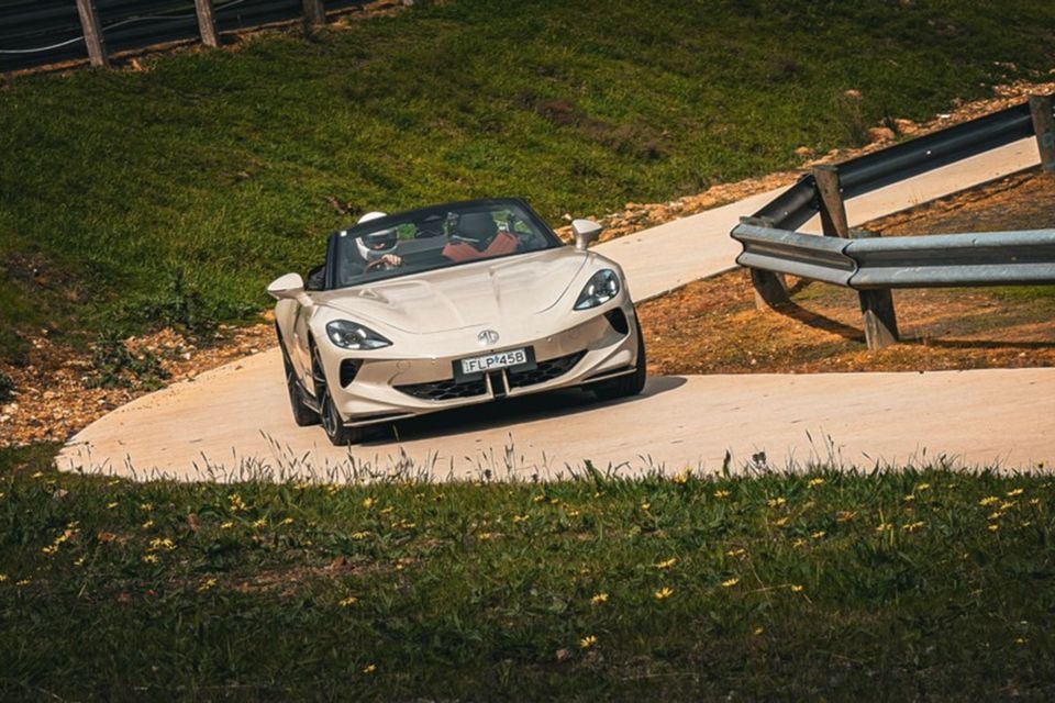 A champagne-coloured MG Cyberster AWD convertible being driven along a winding hillclimb track with steel guardrails, seen from the front
